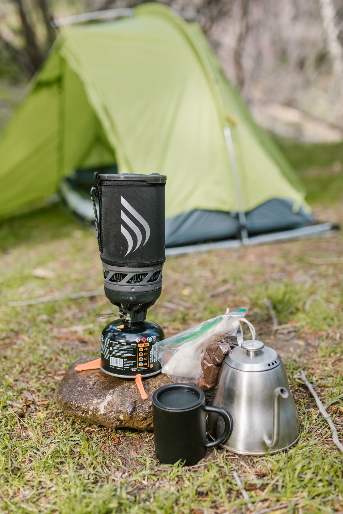 Dog resting on a mat near a camp kitchen setup while someone cooks at a forest campsite