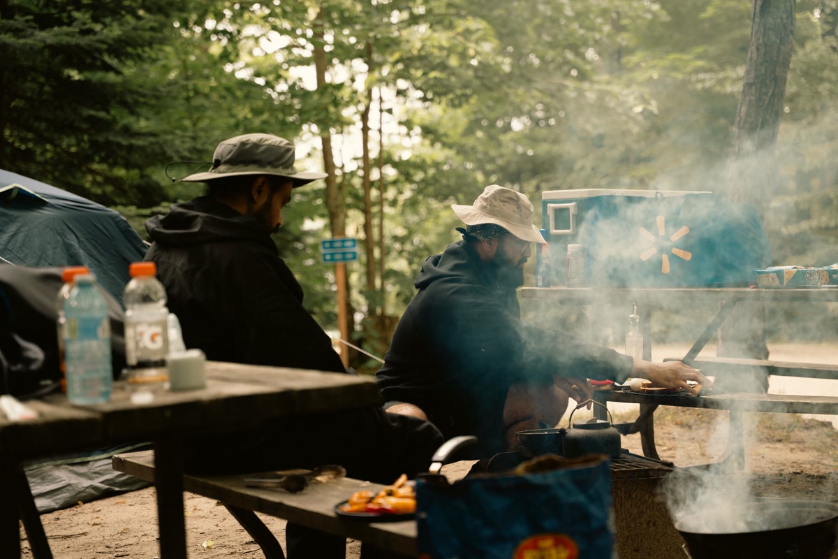 Photograph of three camping pillows arranged on a wooden picnic table at a campsite — one inflatable, one foam, and one hybrid — with a backpack and coffee mug nearby