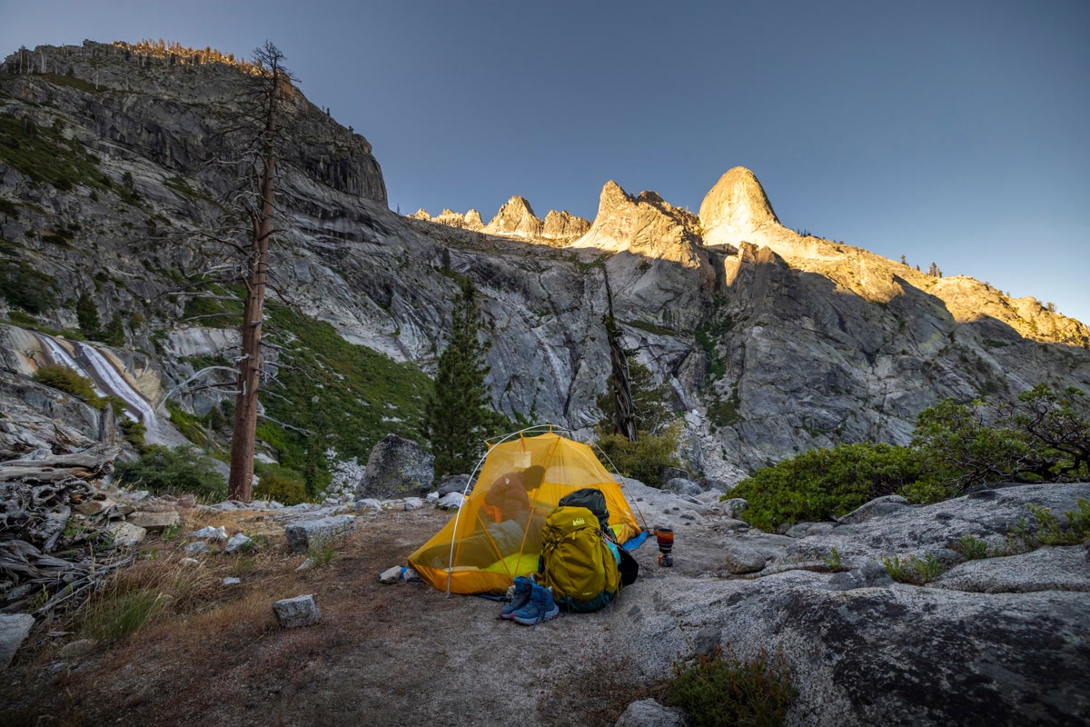 Pair of hiking socks drying on a rock at a backcountry campsite with boots and backpack nearby