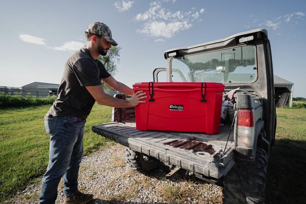 Hard-sided rotomolded cooler with rubber T-latches closed sitting on a truck tailgate at a campsite