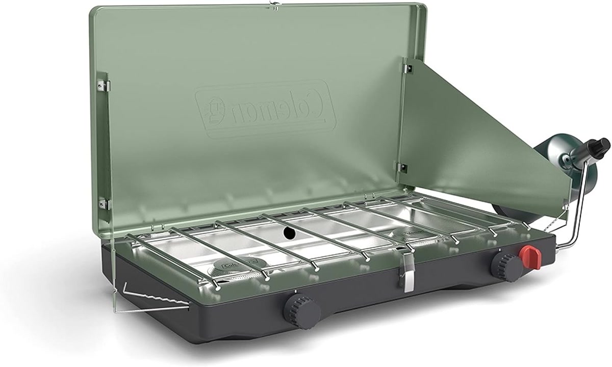 Coleman Cascade Classic stove on a picnic table at a lakeside campsite, showing the wind panels depl