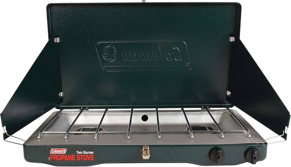 Coleman Classic propane 2-burner stove open on a wooden camp table at dusk, with wind panels up and